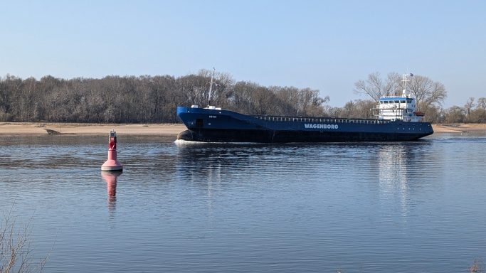 Ein Frachtschiff fährt an einer roten Boje auf ruhigem Wasser vorbei.