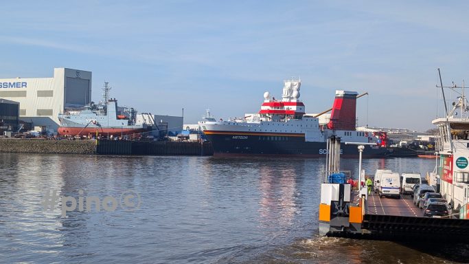 Blick auf mehrere Schiffe am Hafen mit Wasser im Vordergrund und Gebäuden im Hintergrund.