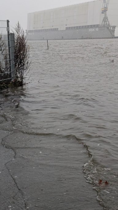 Überschwemmter Bereich mit Wasser, das an einem Zaun und einer großen Wand entlangläuft.