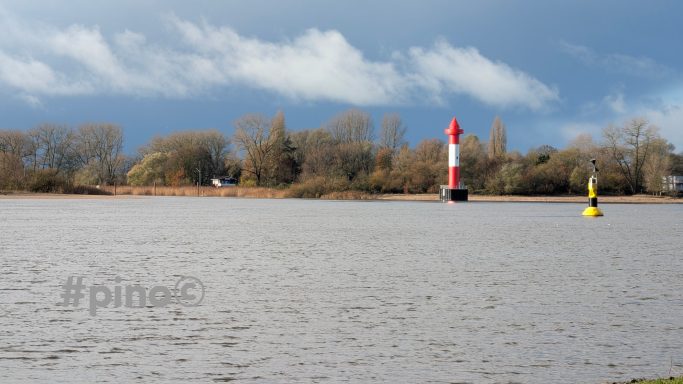 Roter und weißer Leuchtturm neben gelbem Fahrwasserzeichen am Flussufer.