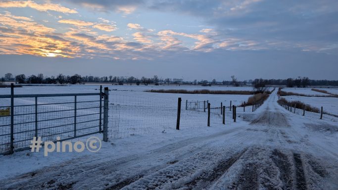 Schneebedeckte Landschaft mit einem Weg, Zaun und Sonnenuntergang im Hintergrund.
