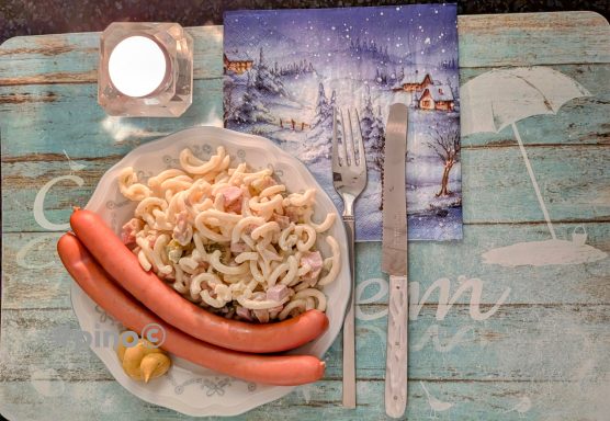 Ein Teller mit Pasta, zwei Würstchen, einem kleinen Glas und einer Serviette auf Holzunterlage.
