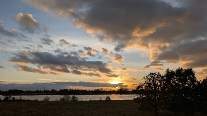Sonnenuntergang über einem Gewässer mit Wolken und silhouettierten Bäumen.