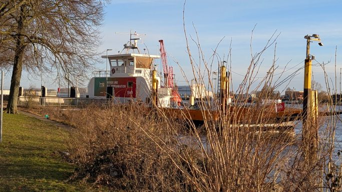 Fähre am Ufer, umgeben von Bäumen und Sträuchern, bei klarem Wetter.