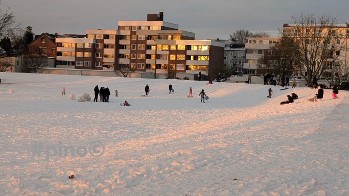 Menschen spielen im Schnee und bauen Schneebälle vor Wohngebäuden bei Sonnenuntergang.