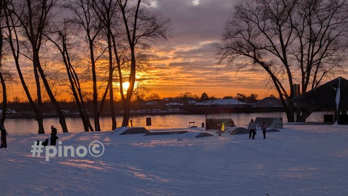 Sonnenuntergang über einem verschneiten Fluss mit Bäumen im Vordergrund.