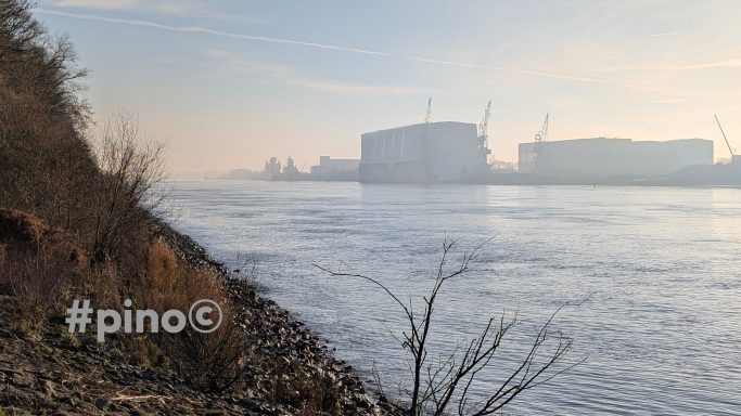 Ruhige Flusslandschaft mit nebligen Ufern und Gebäuden im Hintergrund.