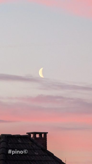 Eine schmale Mondsichel über einem Dach mit pinkem Himmel im Hintergrund.
