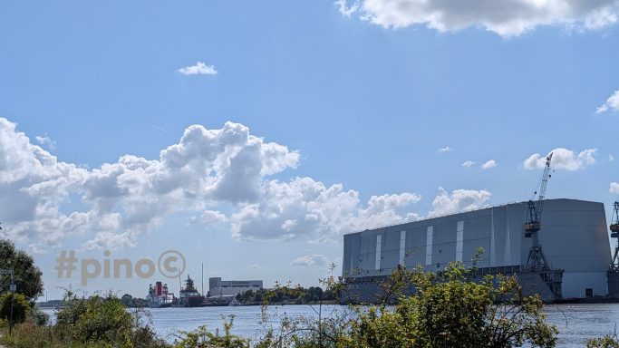 Hafenansicht mit großen Lagergebäuden und einem Schiff am Ufer unter einem blauen Himmel.