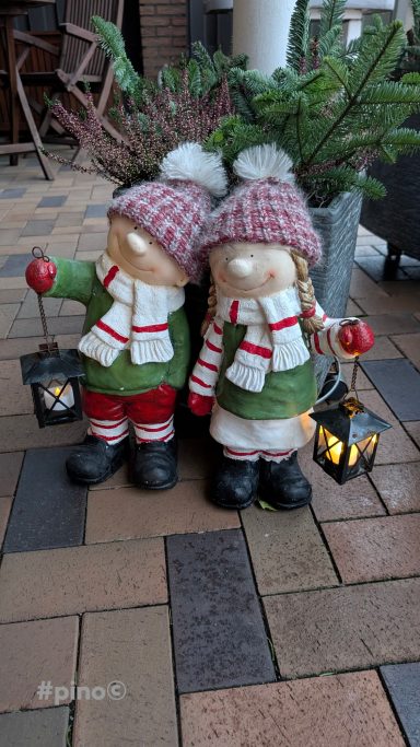 Zwei festlich gekleidete Figuren, die Laternen halten, stehen auf einem Pflasterboden.