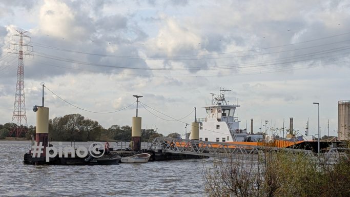 Fährboot am Ufer mit einem großen Schiff im Hintergrund und einer bewölkten Himmel.