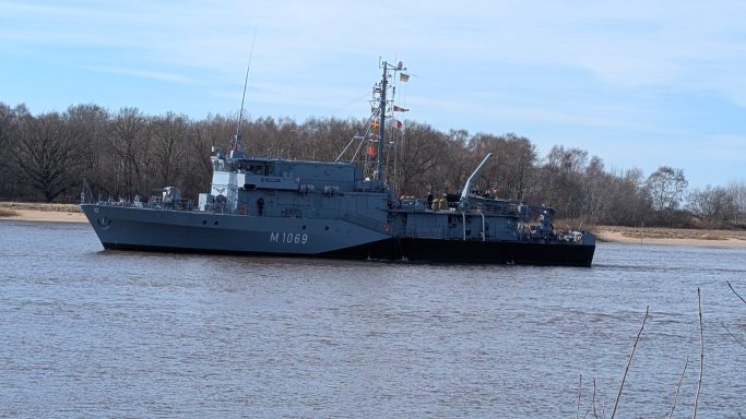 Graues Schiff auf dem Wasser, umgeben von Bäumen und klarem Himmel.