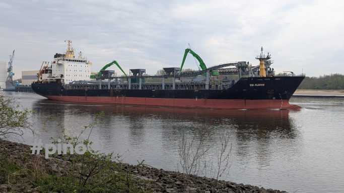 Ein Frachtschiff liegt am Ufer eines Flusses mit Kränen an Deck.