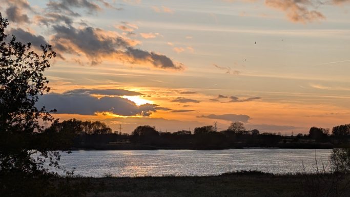 Sonnenuntergang über einem ruhigen See, umgeben von Bäumen und Bewölkung.
