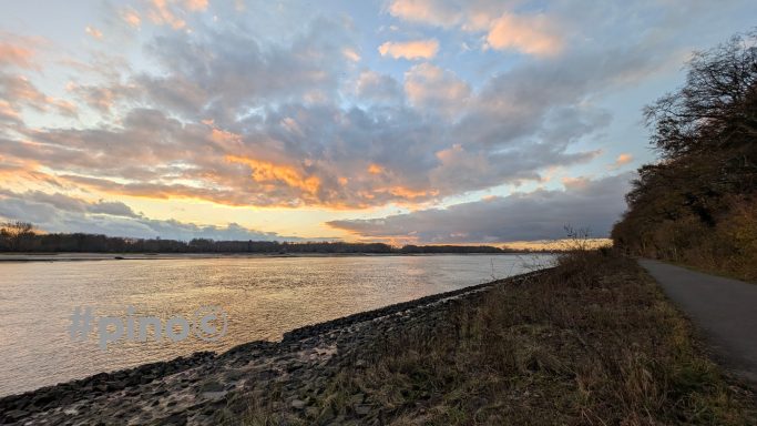 Sonnenuntergang am Fluss mit bunten Wolken und einem schmalen Uferweg.