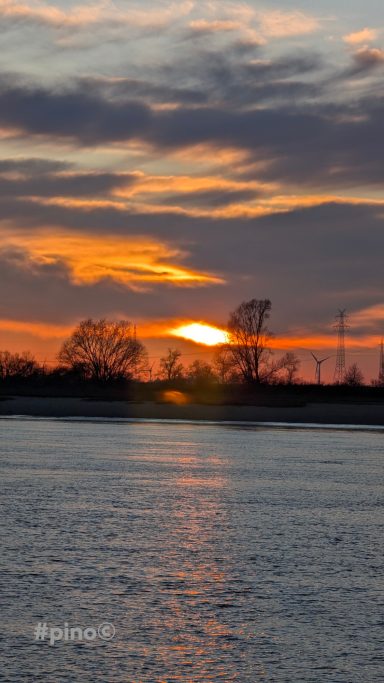 Sonnenuntergang über einem ruhigen Gewässer, umgeben von Bäumen und Wolken.