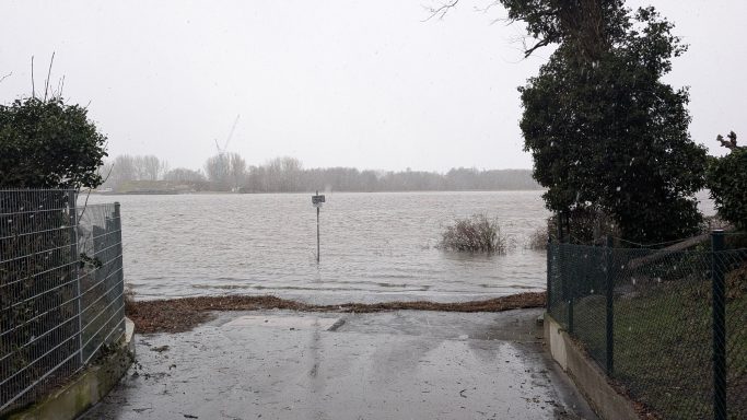 Überfluteter Bereich mit stehendem Wasser und Sicht auf das Ufer. Grauer Himmel.