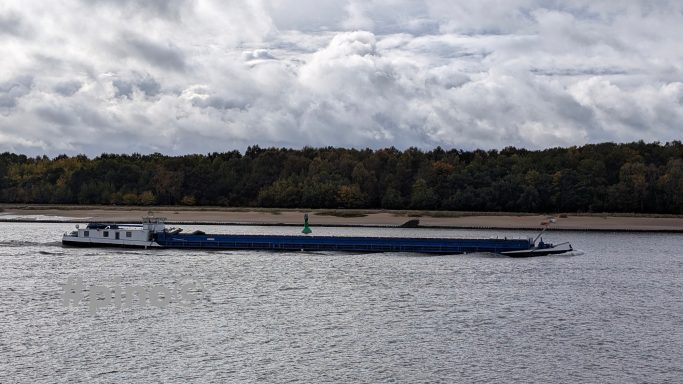 Ein Frachtschiff auf ruhigem Wasser, umgeben von bewaldetem Ufer und bewölktem Himmel.