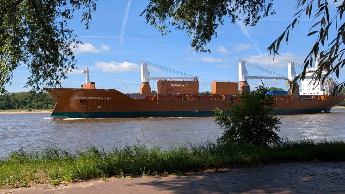 Großes Frachtschiff fährt auf einem ruhigen Fluss unter blauem Himmel.