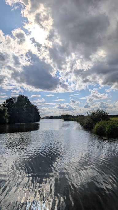 Blick auf einen ruhigen Fluss mit Wolken und Bäumen am Ufer.
