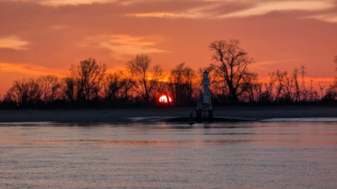 Sonnenuntergang über einem ruhigen Gewässer mit silhouettenhaften Bäumen im Hintergrund.