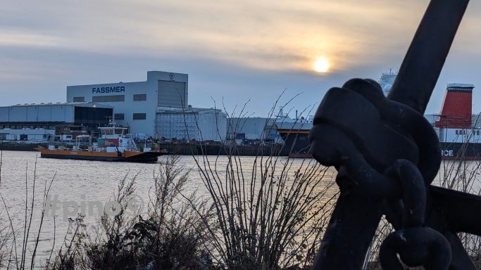 Hafenansicht mit Anker im Vordergrund und Schiff vor der untergehenden Sonne.