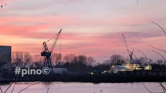 Hafenlandschaft bei Sonnenuntergang mit Kränen und einem Schiff im Hintergrund.