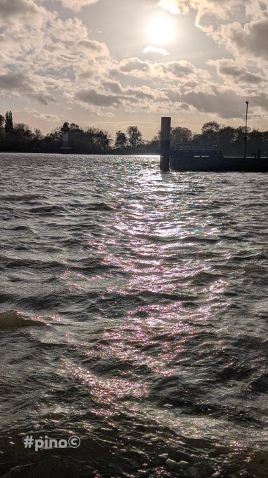 Schimmernde Wasseroberfläche unter einem bewölkten Himmel mit einem Leuchtturm im Hintergrund.