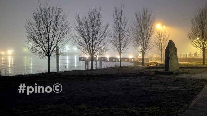 Nebeliger Park mit Bäumen, einem Steindenkmal und beleuchtetem Wasser.