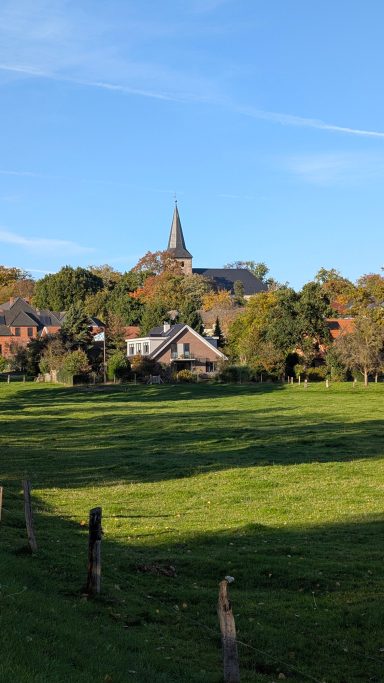 Landschaft mit einem Dorf, einer Kirchturmspitze und grünen Wiesen unter blauem Himmel.