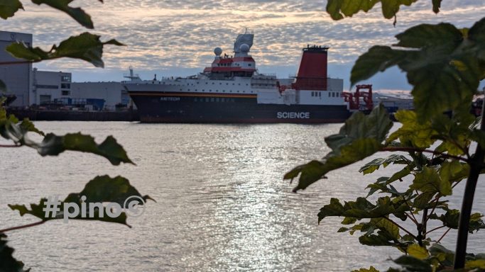 Ein Schiff im Hafen, umgeben von Wasser und Bäumen, unter einem bewölkten Himmel.