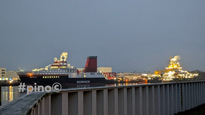 Zwei große Schiffe im Hafen bei Dämmerung, mit Lichtern und reflektierendem Wasser.