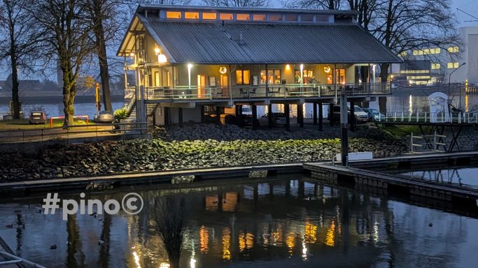 Holzhaus am Wasser bei Dämmerung, umgeben von Bäumen und reflektierendem Wasser.