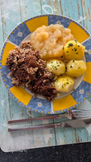 Kartoffeln, Apfelmus und gebratenes Fleisch auf einem bunten Teller.