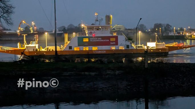 Fähre bei Dämmerung, mit Beleuchtung, am Ufer eines Gewässers.