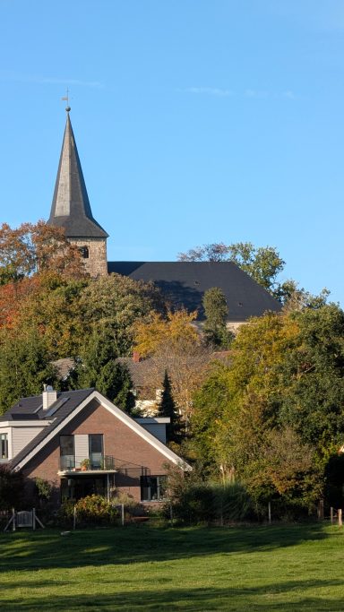 Kirche mit spitzem Turm über einem Dorf, umgeben von Herbstbäumen.