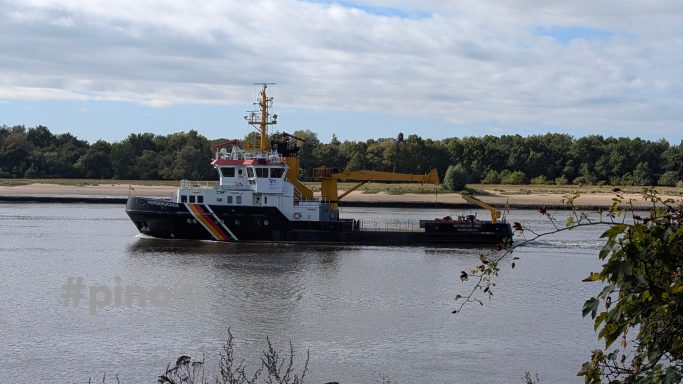 Ein Schlepper fährt auf einem Fluss mit grüner Ufervegetation im Hintergrund.