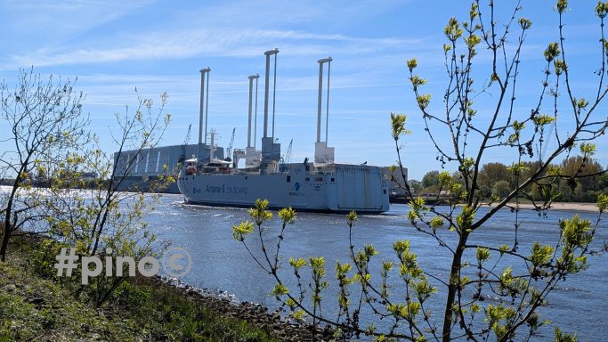 Ein Schiff fährt an einem Kraftwerk mit hohen Schornsteinen entlang, umgeben von Bäumen.