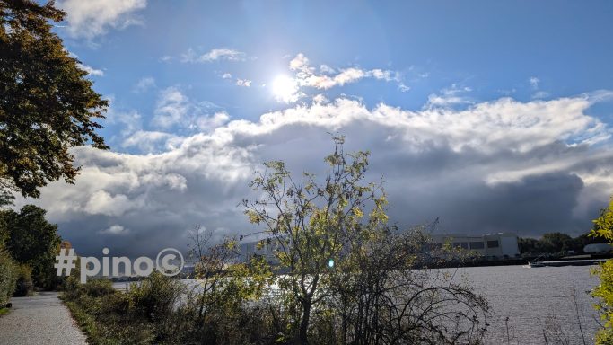 Himmel mit Wolken und Sonne über einem ruhigen Gewässer, umgeben von Bäumen.