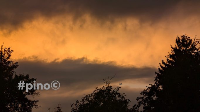 Dramatischer Himmel mit orangefarbenem Licht und dunklen Wolken, silhouettierte Bäume.