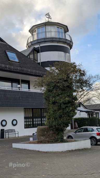 Leuchtturm neben einem modernen Gebäude, umgeben von Bäumen und einem Parkplatz.