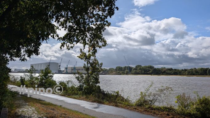 Ein Fluss mit Ufervegetation und Wolken am Himmel. In der Ferne sind Gebäude sichtbar.