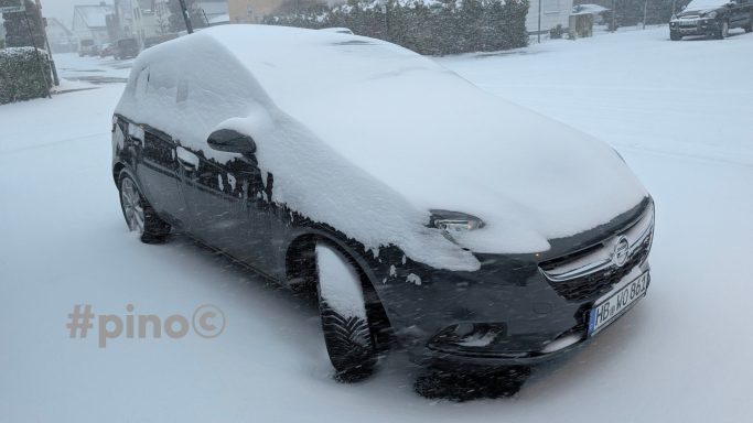 Schwarzes Auto, komplett mit Schnee bedeckt, in winterlicher Landschaft.