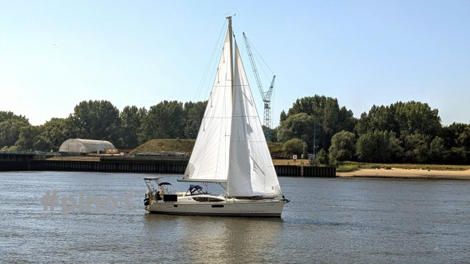 Ein Segelboot mit weißen Segeln fährt auf ruhigem Wasser bei sonnigem Wetter.