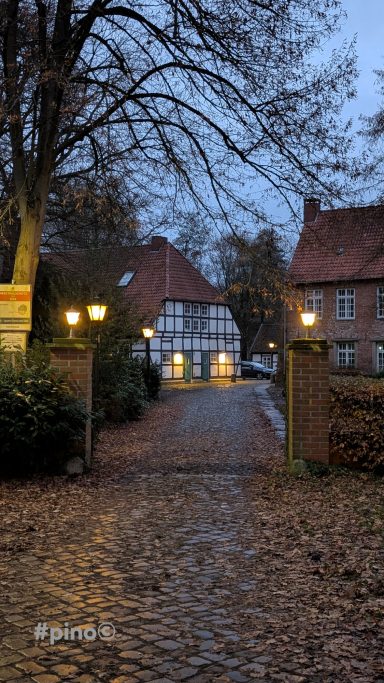 Eingang zu einem Fachwerkhaus, umgeben von Bäumen und Laternen bei Dämmerung.