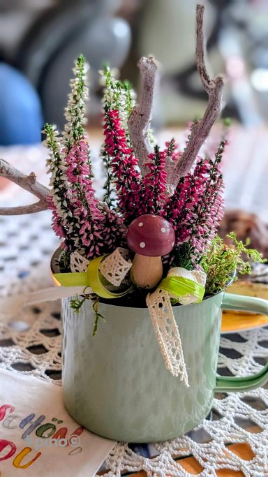 Topf mit bunten Heidepflanzen, Kiefernnadeln und einem kleinen Pilz.