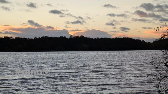 Ruhiger See bei Sonnenuntergang, mit Wolken und einer bewaldeten Uferlinie.