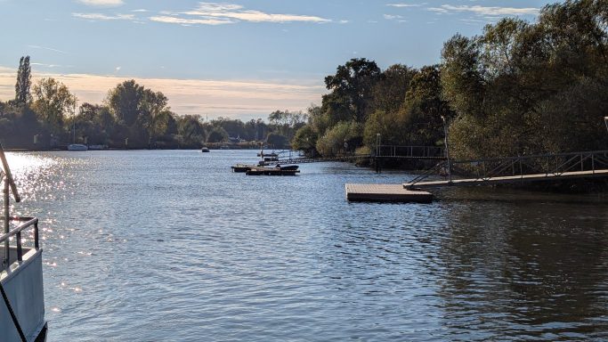 Ruhiger Fluss mit Boot und einer niedrigen Holzplattform am Ufer, umgeben von Bäumen.