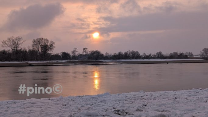Sonnenuntergang über einem verschneiten See, mit reflektierendem Licht auf dem Wasser.