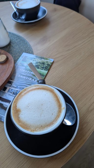 Schwarzes Kaffeetasse mit Milchschaum auf einem Holztisch, daneben eine kleine Gabel.